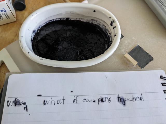 A plastic tub of black iron-gall ink, along with handwriting on a lined notebook using this ink, showing pale blue-black lines and splotches of ink splatter.